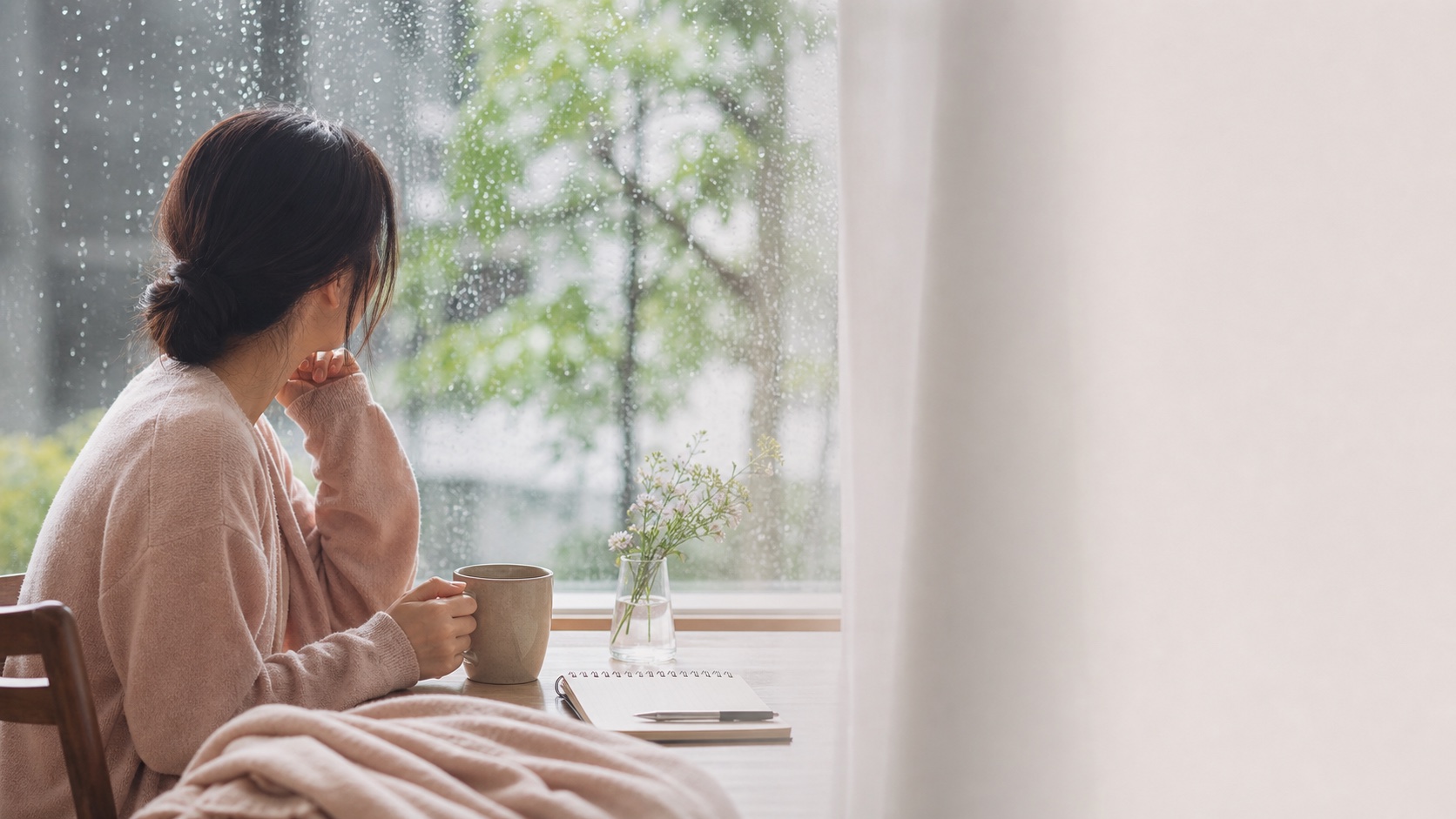 雨の降る窓辺で温かい飲み物を持ち、静かに休む女性の横姿